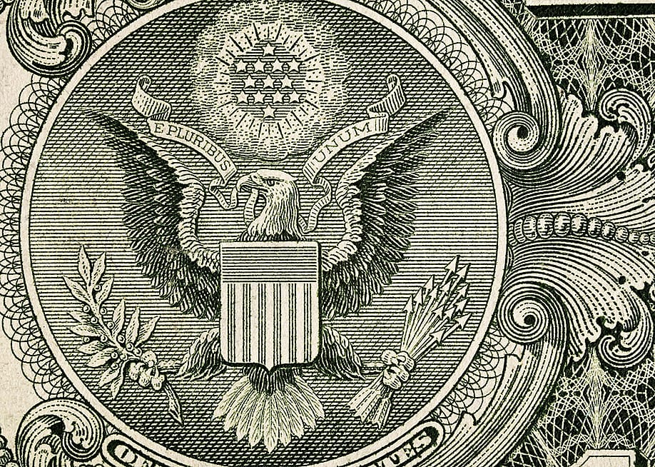 A close-up shot showing the macro details with the engraved eagle, shield, stars, and ornamental line work of the Great Seal of the United States on the one-dollar banknote. — Yulcha // Shutterstock