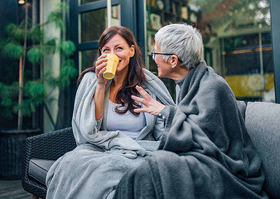 Two people wrapped in cozy throw blankets while sitting outdoors and sharing a warm drink. — Branislav Nenin // Shutterstock