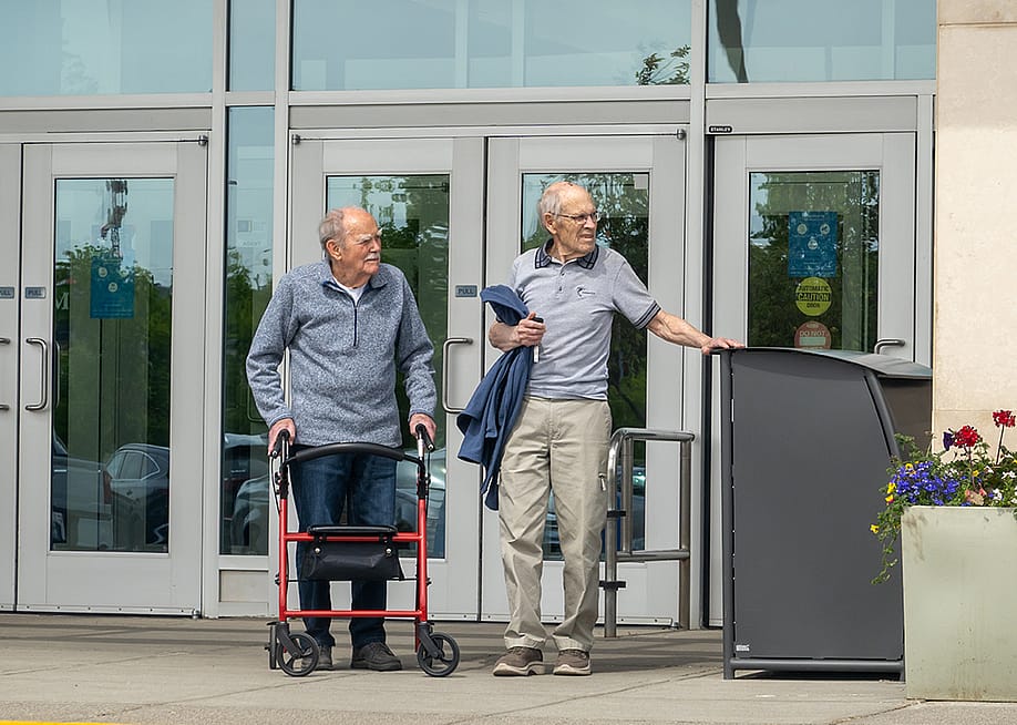 Two elderly men talking outside a building entrance where one is using a red rollator walker. — Habanero Pixel // Shutterstock