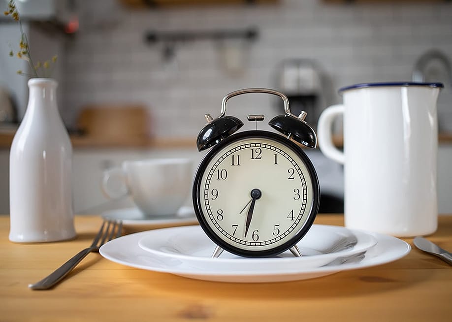 An alarm clock placed in a plate on the kitchen table, representing an intermittent fasting concept. — Melinda Nagy // Shutterstock