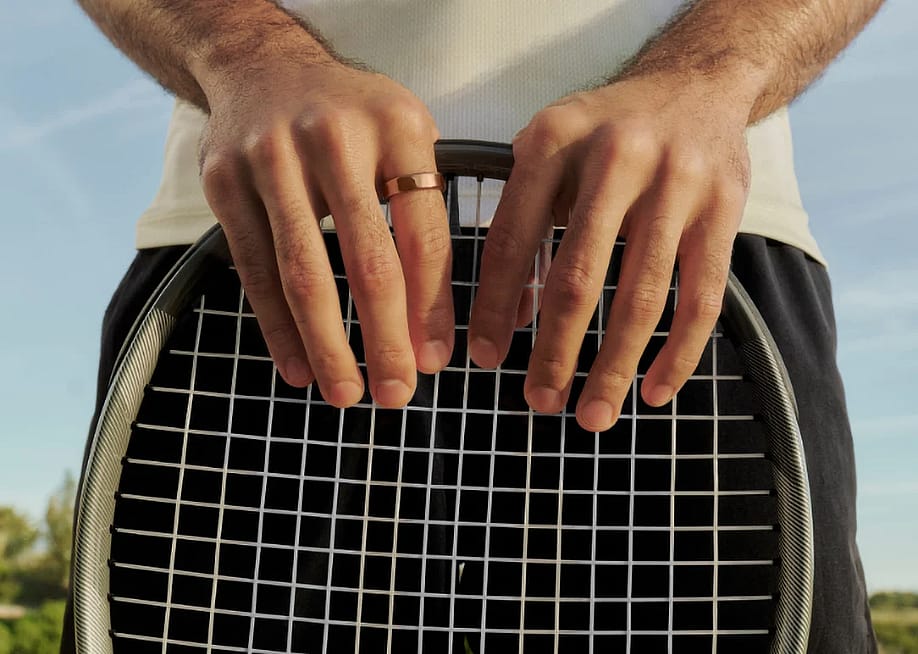 A person wearing a smart ring on his finger and holding a tennis racket outdoors. — Oura