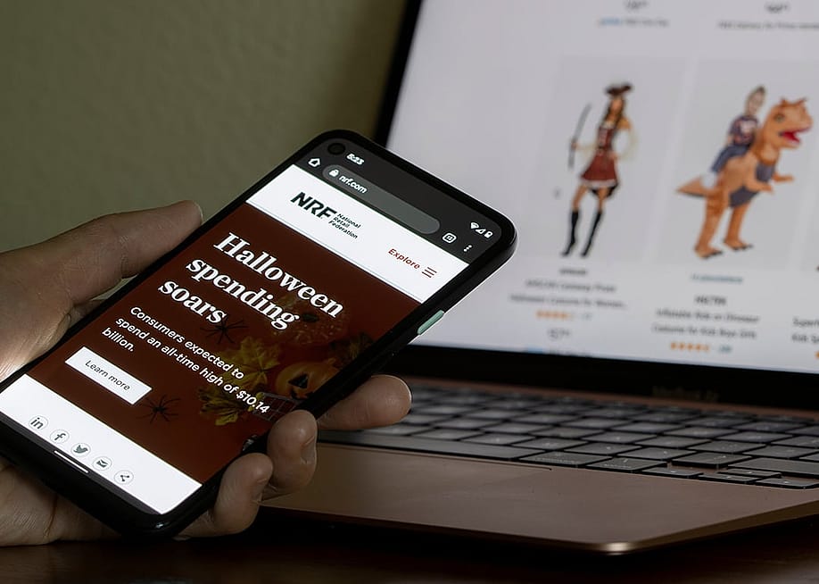 A person reading an article about Halloween spending forecast from the National Retail Federation (NRF) website on her smartphone with an online Halloween store in the background. — Tada Images // Shutterstock