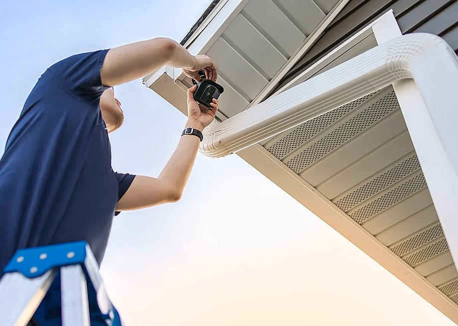 A technician on ladder installing a wireless security video camera underneath a private house's roof for its outdoor surveillance system. — ANNVIPS // Shutterstock