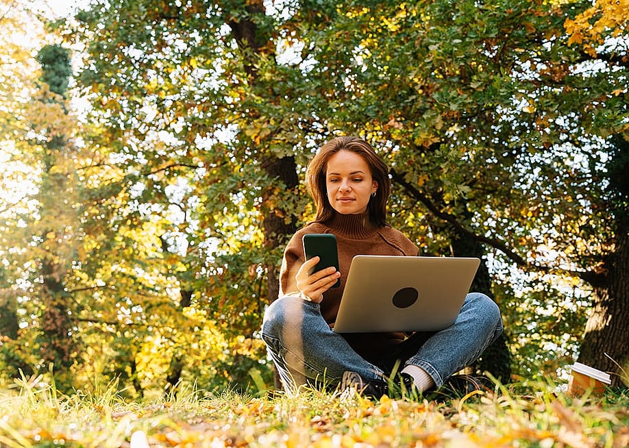 A young adult working outdoors on a laptop and her smartphone in a park. — Igor Barilo // Shutterstock
