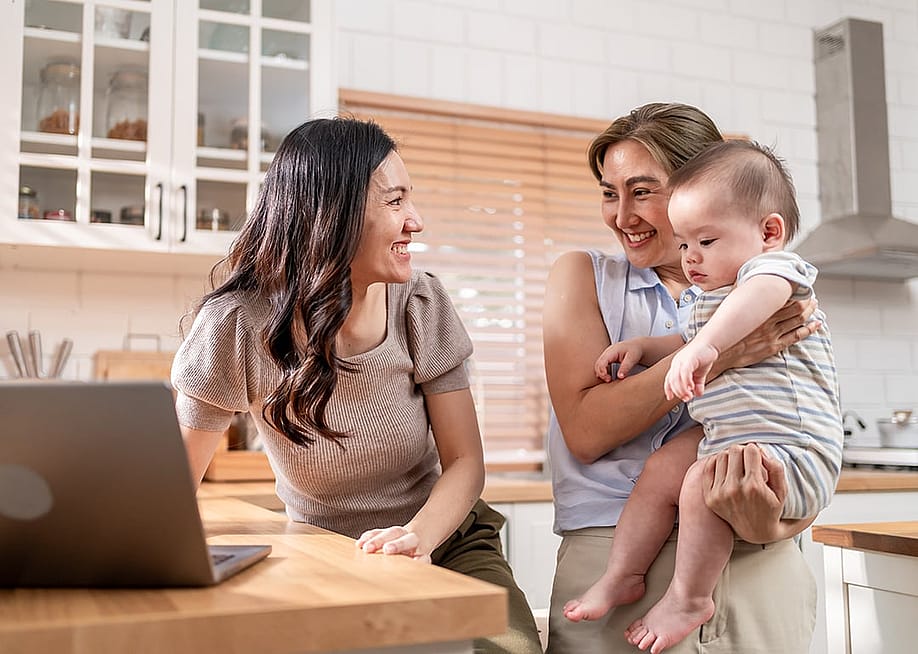 An elder sibling cradling a baby while the other sibling working remotely from their kitchen area. — Creation Company // Shutterstock