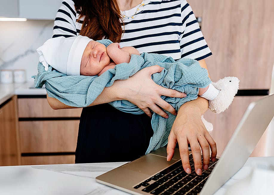 A parent cradling a newborn baby while working remotely from their home kitchen. — Iryna Inshyna // Shutterstock
