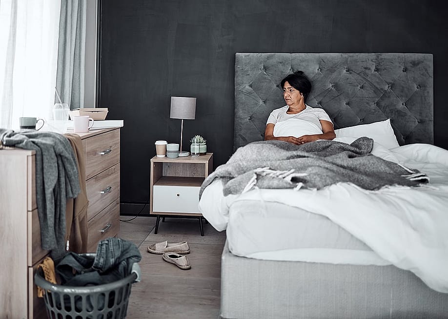 An elderly person feeling sad and zoning out in her unorganized bedroom alone. — PeopleImages // Shutterstock