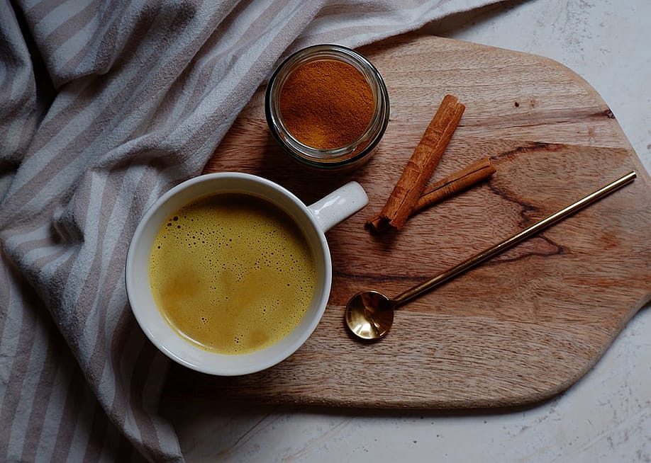 A cup of hot turmeric latte with cinnamon powder and sticks placed on a wooden cutting board. — re_bekka // Shuttterstock