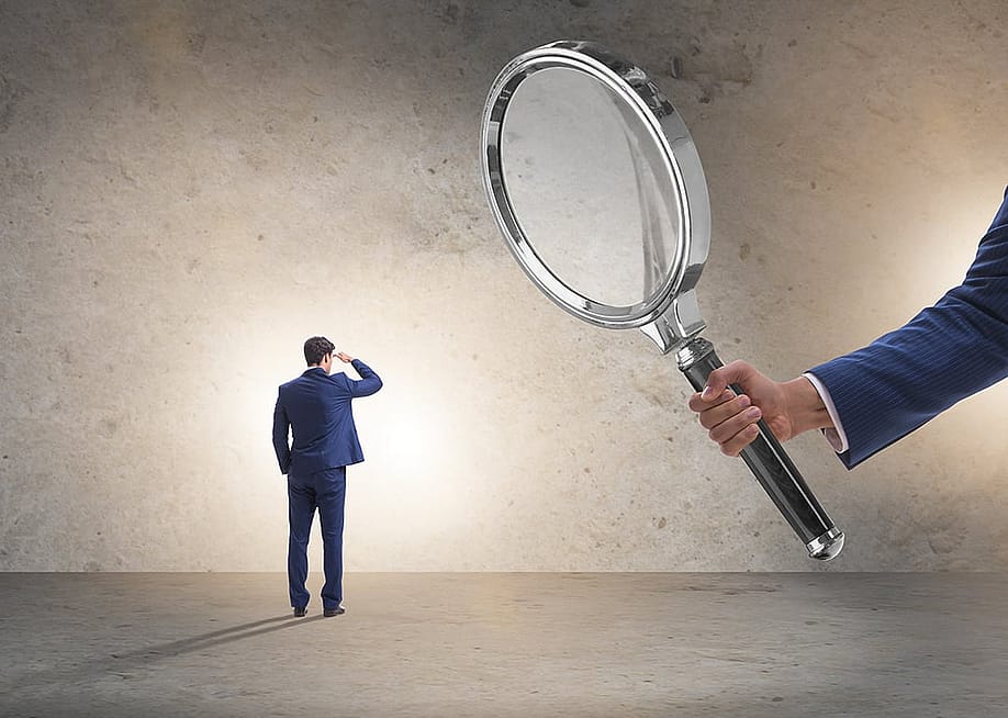 A giant hand holding a huge magnifying glass over a miniature employee, representing work monitoring or surveillance concept. — Elnur // Shutterstock
