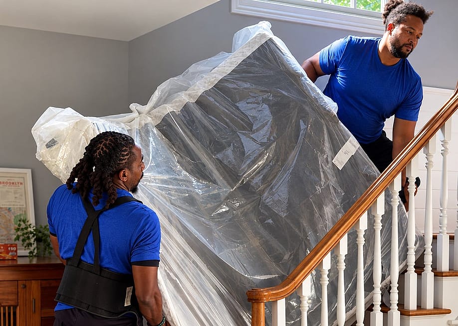 Professional movers carrying a plastic-wrapped mattress upstairs inside a home. — Lugg