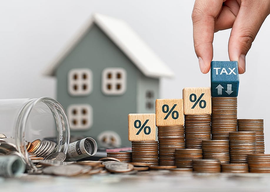 Stacks of coins, wooden blocks interest and tax symbols, and a house model seen in the background, representing property taxes. — dee karen // Shutterstock