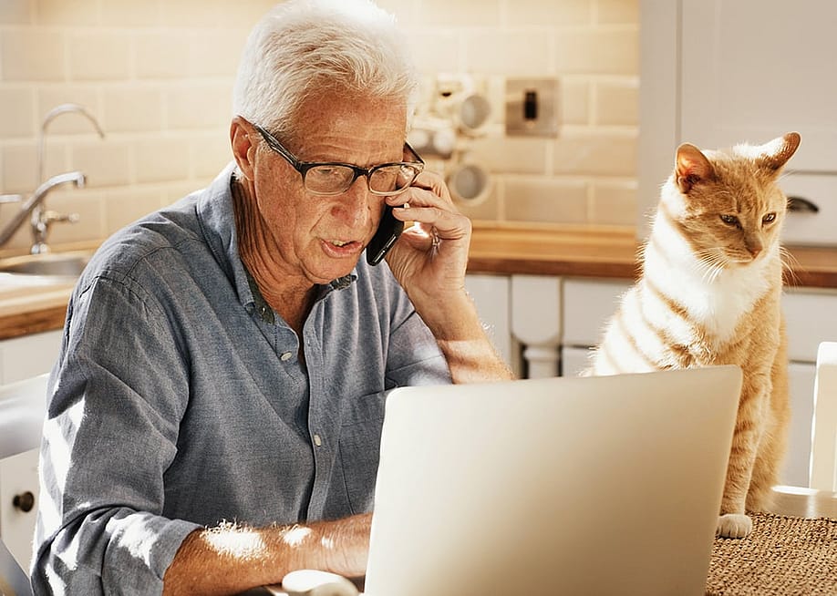 An elderly person talking to an insurance officer over the phone while checking his files on a laptop in the dining table. — PeopleImages // Shutterstock