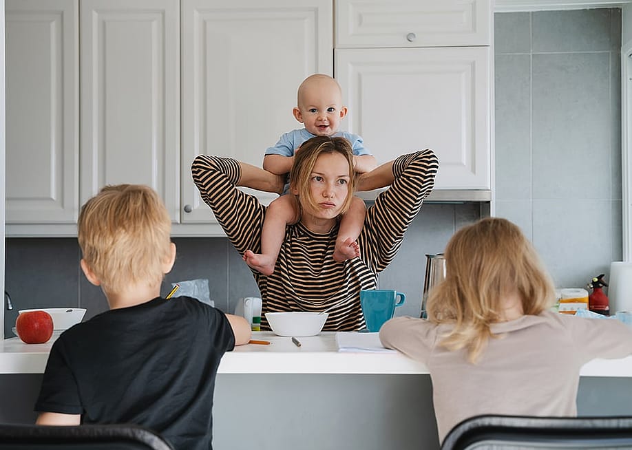 A parent carrying her baby on her shoulders with her other children doing school homeworks in the kitchen countertop. — Natalia Deriabina // Shutterstock