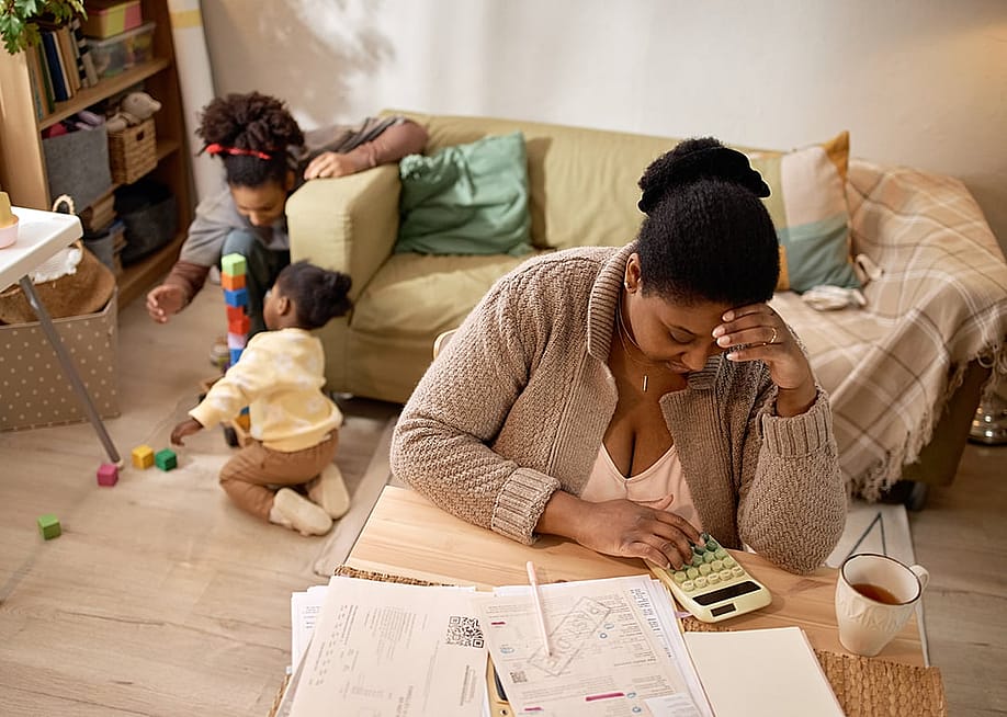 A parent calculating their family's budget, savings, and expenses while children playing in the living area. — SeventyFour // Shutterstock