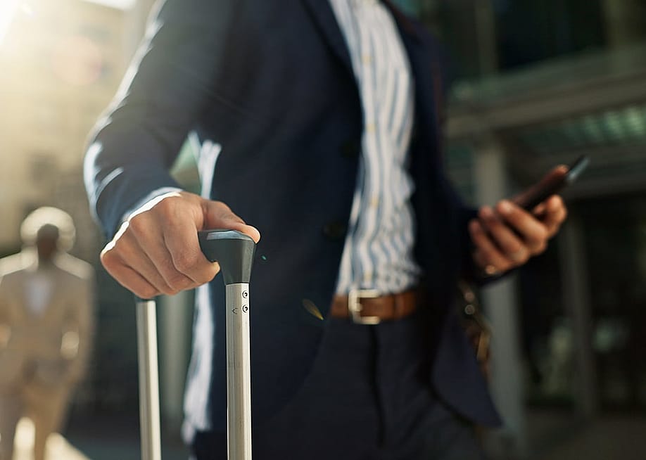 A business professional holding a smartphone and suitcase while navigating a specific location outside the airport. — PeopleImages // Shutterstock