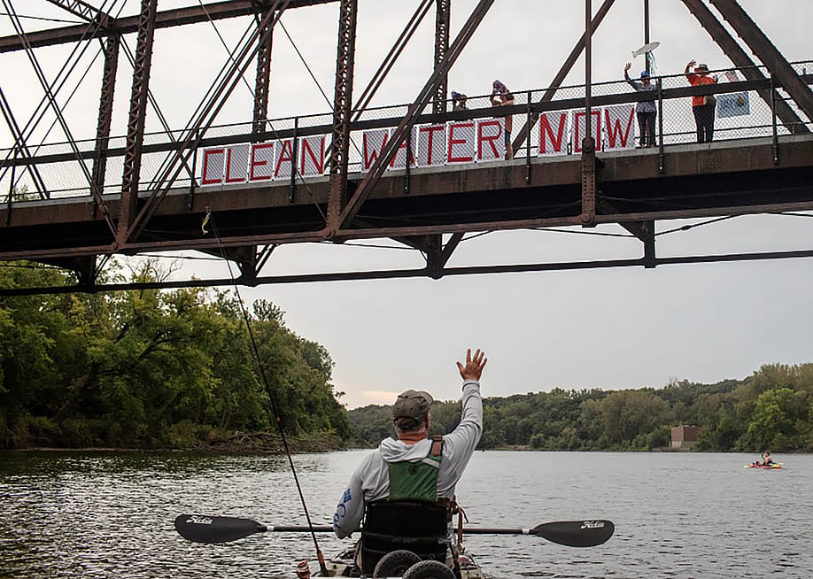 Chris Jones, in a kayak on the river, waves to supporters demonstrating on a bridge for clean water in Des Moines, Iowa. Jones, a retired scientist and water advocate, is running for Iowa’s secretary of agriculture. — Danny Wilcox Frazier