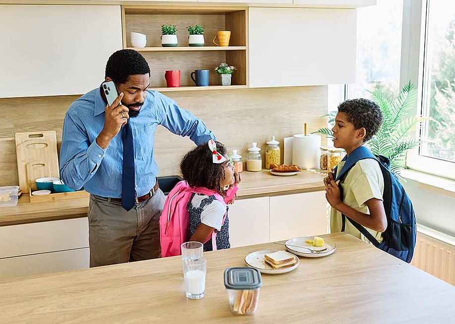 A father getting his son and daughter ready for school while also taking a call on the phone. — pics five // Shutterstock