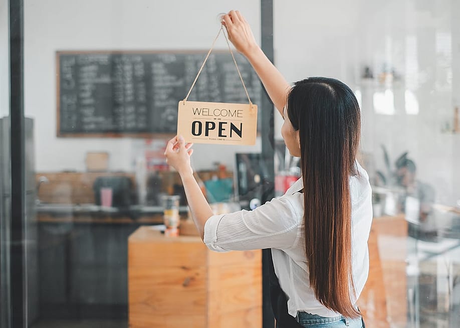 A cafe owner happily puts up a 'Welcome, we are open' sign by the entrance of her shop. — Mongta Studio // Shutterstock