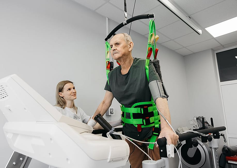 Elderly man undergoing verticalization therapy with support from a female therapist in a well-equipped rehabilitation center featuring therapy machines. — wedmoments.stock // Shutterstock