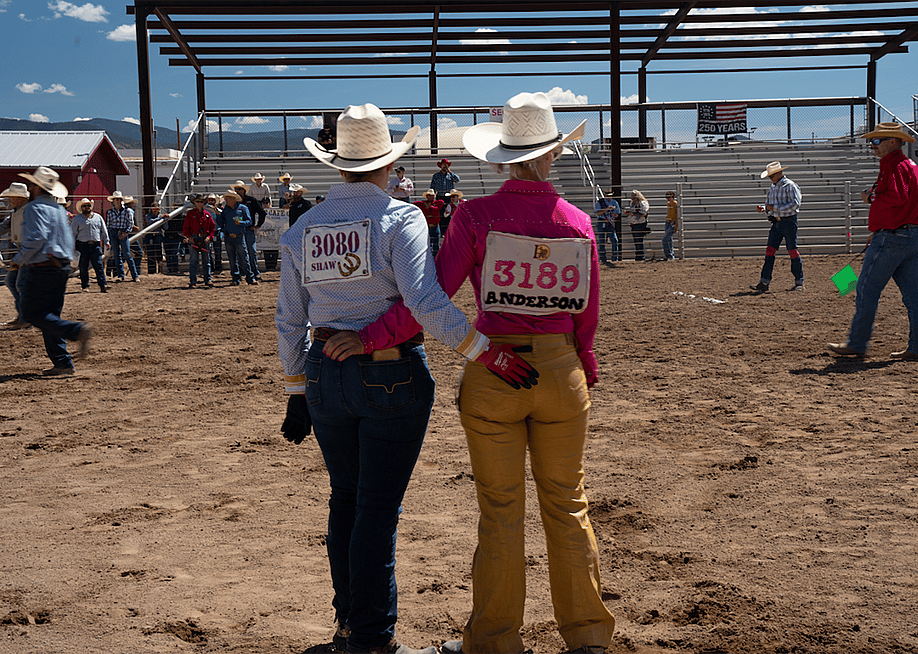 Two rodeo participants stand with their arms around each other's backs looking at the rodeo grounds. — Anya Petrone Slepyan // The Daily Yonder