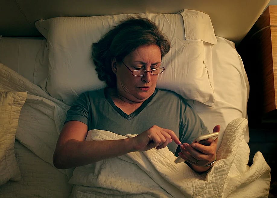 A woman lying in bed under a white blanket, scrolling or interacting with a smartphone. — Bricolage // Shutterstock