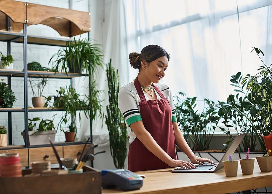 A smiling female entrepreneur wearing an apron works on laptop in her plant store, surrounded by greenery. — LightField Studios // Shutterstock