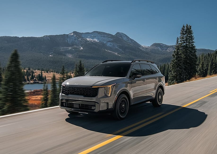 Tan 2025 Kia Sorento driving on a road with mountains and a lake in the background. — Kia