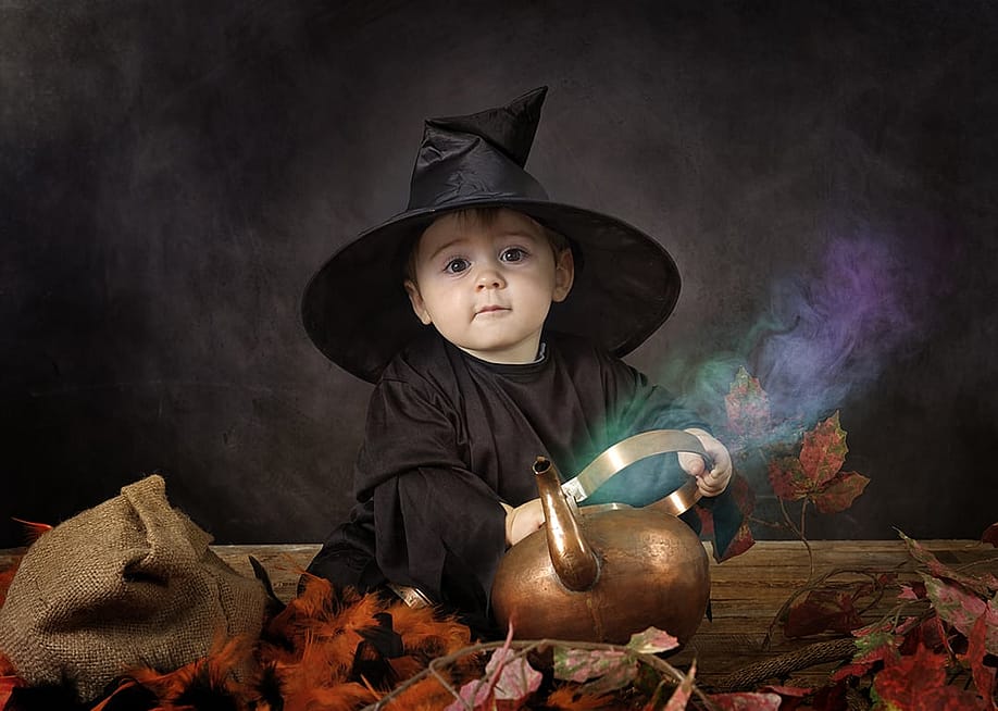A young baby dressed in a classic black witch costume with a pointed witch hat, sitting beside a copper pot or kettle with colorful smoke or steam rising from it. — aldegonde // Shutterstock