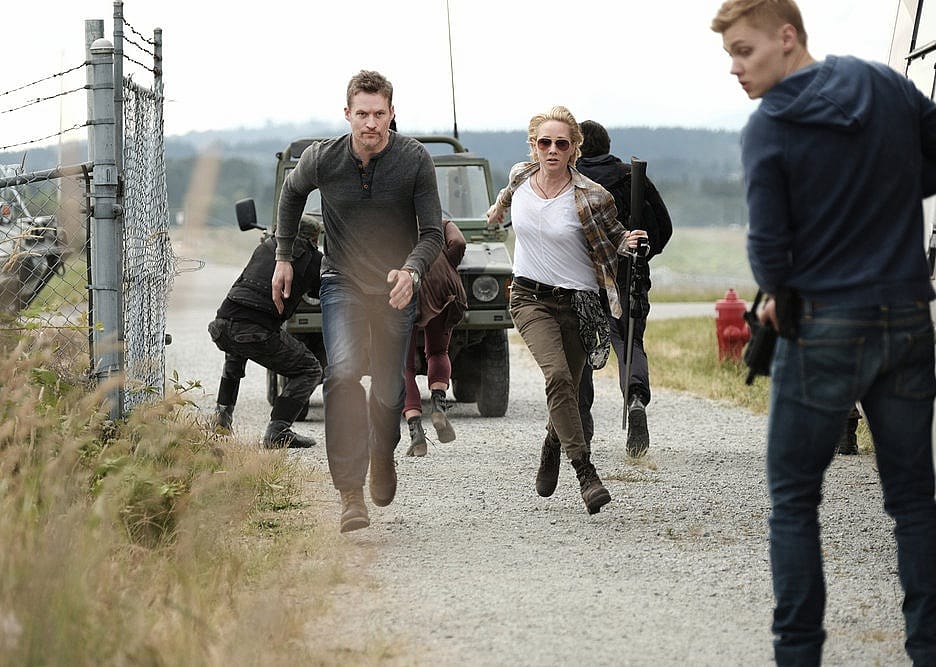 A man and a woman run through a gate from a moving army jeep while others try to cover for them with weapons.