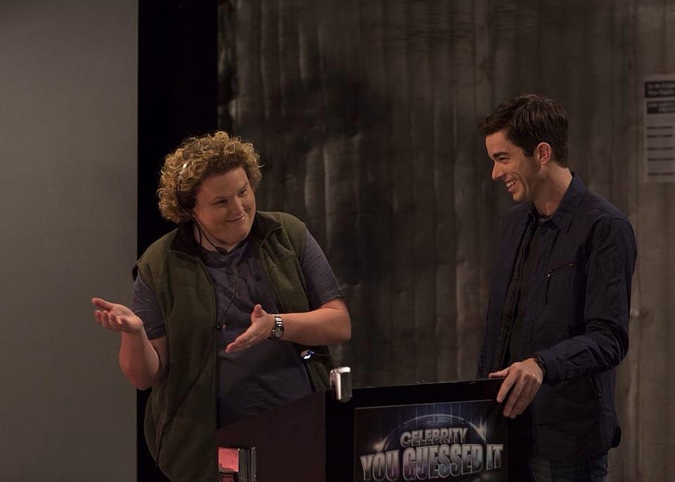 A man stands behind a game show podium laughing at a crew member.
