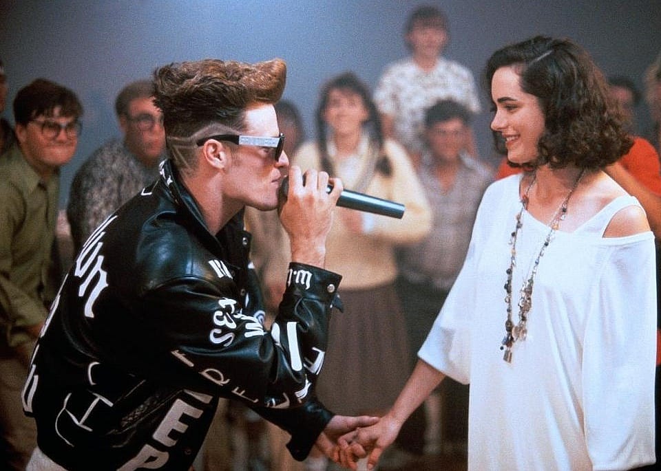 Vanilla Ice in a black and white leather jacket singing to a woman in a white off the shoulder shirt.